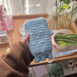 Op de afbeelding: Een lichtblauwe gehaakte walvisvormige etui met witte hartvormige accenten. De etui heeft kleine zwarte ogen en een wit trekkoord. De walvis heeft kleine vinnen en een gerimpelde staart. De etui is handgemaakt.