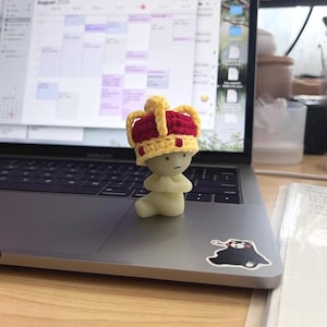 Op de afbeelding: Een klein, wit beeldje dat een gele en rode gebreide kroon draagt. Het beeldje staat in gebedshouding en staat op een zilveren laptop.