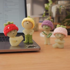 May include: Four small crocheted figures with different colored hats. The figures are made of a light-colored material and have simple facial features. The hats are red, yellow, purple, and pink.