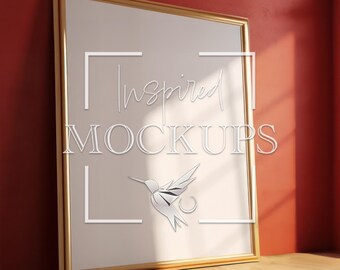 May include: A gold-framed poster mockup with a white background. The poster is leaning against a red wall and a wooden floor. The poster has a white hummingbird graphic and the text "Inspired Mockups" in the center.