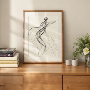 May include: A black and white line drawing of a person in a flowing dress, framed in a light wood frame. The artwork is displayed on a wooden shelf with books and plants.