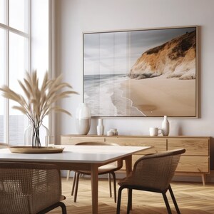 Puede incluir: Una mesa de comedor blanca con un marco de madera clara y cuatro sillas tejidas. La mesa est&aacute; puesta con una bandeja dorada y un jarr&oacute;n de hierba de la pampa seca. Una gran impresi&oacute;n enmarcada de una escena de playa cuelga en la pared sobre un aparador de madera.