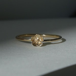 May include: A delicate gold ring featuring a small, textured flower design. The ring's band is thin and polished, and the flower is the focal point. The ring is set against a neutral background, highlighting its elegant simplicity.