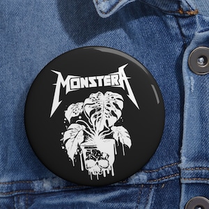 May include: Black and white button with a skull-shaped pot with a Monstera plant growing out of it. The text "MONSTERA" is above the plant in a metal band style font.