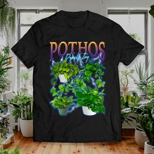 Op de afbeelding: Zwart T-shirt met een afbeelding van Pothos-planten, ook bekend als duivels-klimop, met een bliksemontwerp. Het woord "Pothos" staat in oranje en paarse tekst. Het shirt toont verschillende planten in potten.