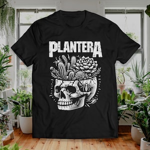 Op de afbeelding: Zwart t-shirt met een witte afbeelding van een schedel met planten die eruit groeien. Het woord "PLANTERA" is in een verweerde lettertype boven de schedel gedrukt. Het ontwerp bevat cactussen, vetplanten en een insect.