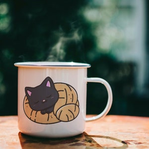 Puede incluir: Taza de esmalte blanco con un gato negro acurrucado dentro de un pastel marr&oacute;n. La taza tiene un borde y un asa azules. Sale vapor de la taza.