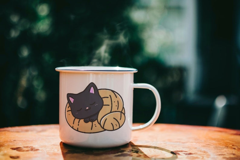 Puede incluir: Taza de esmalte blanco con un gato negro acurrucado dentro de un pastel marr&oacute;n. La taza tiene un borde y un asa azules. Sale vapor de la taza.