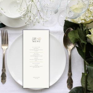 Peut inclure: Une carte de menu blanche avec des lettres dorées et un petit motif floral. La carte de menu indique "The Menu" et liste l'entrée, le plat principal, le dessert et les boissons.