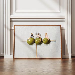 May include: A framed art print depicting three large green olives, each with a small figure seated on top. The artwork is set against a textured white background and framed in a brown wooden frame. The print is displayed on a wooden floor.