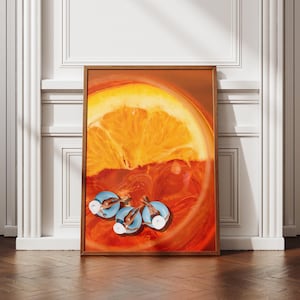 May include: A framed art print showing a close-up of an orange slice in a reddish-orange liquid, with three small figures swimming in the liquid. The print is displayed on a wooden floor against a white wall.