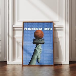 May include: A framed rectangular poster featuring a blue background with the repeating white text IN KNICKS WE TRUST. The central graphic depicts a green statue hand holding a torch, with an orange basketball balanced on top of the torch flame.