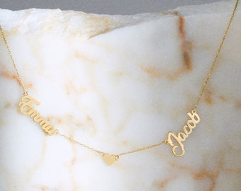 May include: Gold necklace with two names, "Emma" and "Jacob", connected by a small heart charm. The necklace is displayed on a white marble surface.
