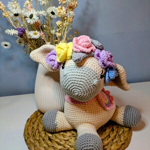 Könnte beinhalten: Handgefertigte Häkel-Einhorn-Puppe mit grauem Kopf, cremefarbenem Körper und grauen Hufen. Das Einhorn hat einen rosa Schal und einen Blumenkranz mit gelben, rosa und lila Blumen. Es sitzt auf einer gewebten Matte mit einer Vase mit Trockenblumen im Hintergrund.