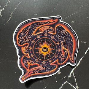 May include: A sticker with a black and orange design of a winged eye surrounded by a circle of smaller eyes. The eye is in the center of a sunburst design.