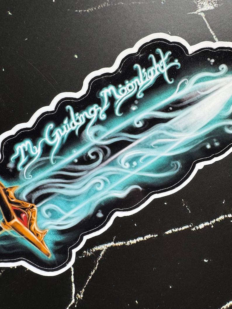 May include: A sticker featuring a blue and white sword design. The sword is surrounded by a swirling, smoky effect. The text "The Guiding Moonlight" is written in a stylized font above the sword.