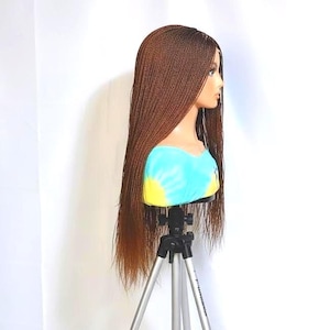 May include: A brown braided wig on a mannequin head. The wig is long and straight, with a natural-looking color gradient. The mannequin is supported by a tripod stand, and the background is plain white.