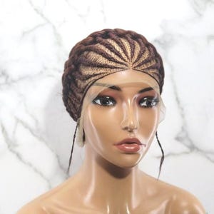 May include: A brown braided wig with a lace front is displayed on a mannequin. The wig features intricate braiding patterns and a natural-looking hairline. The mannequin's skin tone is a warm brown, and the background is a white marble pattern.