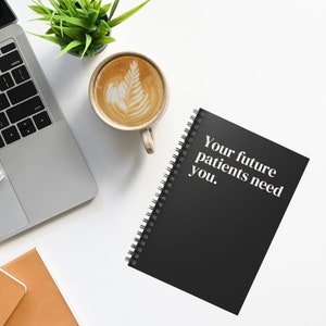 May include: A black spiral bound notebook with the text "Your future patients need you."  The notebook is on a white surface with a laptop, a cup of coffee, and a potted plant.