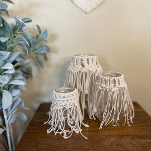 May include: Three glass candle holders with cream-colored macrame covers. The macrame features a geometric pattern at the top and long fringe. The candle holders are arranged on a wooden surface, with a leafy plant in the background.
