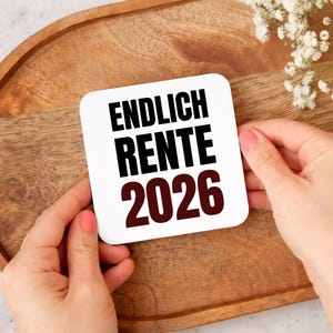 May include: A white square card with the words "ENDLICH RENTE 2026" in black and burgundy. The card is held by two hands with pink nail polish, set against a wooden tray.