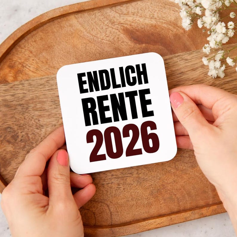 May include: A white square card with the words "ENDLICH RENTE 2026" in black and burgundy. The card is held by two hands with pink nail polish, set against a wooden tray.