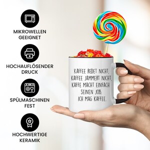 May include: White ceramic coffee mug with a black handle. The mug has black text that reads "Kaffee redet nicht, Kaffee jammert nicht, Kaffe macht einfach seinen Job. Ich mag Kaffee."  The mug is sitting on a hand with a colorful lollipop sticking out of the top.