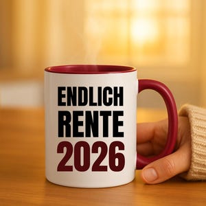 May include: A white ceramic mug with a burgundy interior and handle. The mug features the text "ENDLICH RENTE 2026" in black and burgundy. Steam rises from the mug, suggesting a hot beverage. The mug is held by a person.