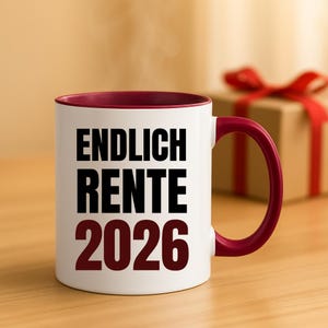 May include: White ceramic mug with a burgundy interior and handle. The mug features the text "ENDLICH RENTE 2026" in bold black and burgundy lettering. A gift box with a red ribbon is in the background.