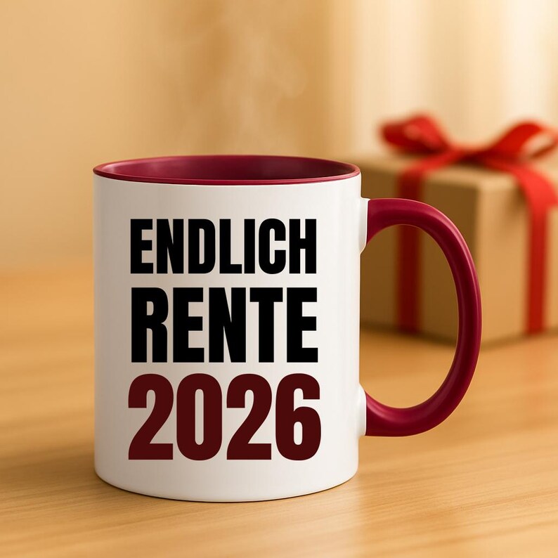 May include: White ceramic mug with a burgundy interior and handle. The mug features the text "ENDLICH RENTE 2026" in bold black and burgundy lettering. A gift box with a red ribbon is in the background.