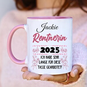Puede incluir: Taza de cer&aacute;mica blanca con asa e interior rosas. La taza presenta el nombre "Jackie" y la palabra "Rentnerin" en escritura rosa, junto con el a&ntilde;o "2025" y una frase en alem&aacute;n.
