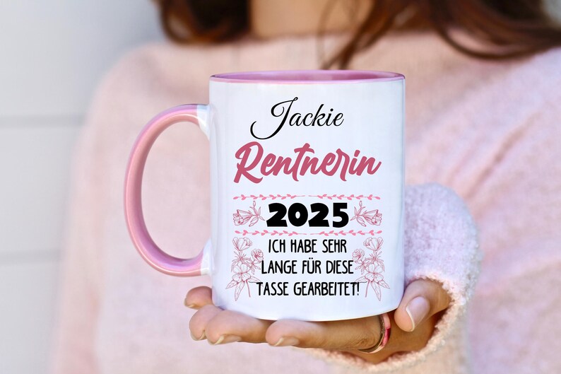 Puede incluir: Taza de cer&aacute;mica blanca con asa e interior rosas. La taza presenta el nombre "Jackie" y la palabra "Rentnerin" en escritura rosa, junto con el a&ntilde;o "2025" y una frase en alem&aacute;n.