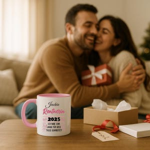Puede incluir: Una taza de cer&aacute;mica blanca con interior y asa rosas. La taza presenta el texto "Jackie Rentnerin 2025" en fuente cursiva, junto con texto adicional en alem&aacute;n. La taza est&aacute; sobre una mesa de madera, con una pareja en el fondo.