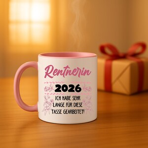 May include: A white ceramic mug with a pink handle and interior. The mug features the German text "Rentnerin 2026" in pink, along with the phrase "Ich habe sehr lange f&uuml;r diese Tasse gearbeitet!" in black, and floral accents.