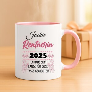 Puede incluir: Taza de cer&aacute;mica blanca con interior y asa rosas. La taza presenta el nombre "Jackie" y la palabra "Rentnerin" en escritura rosa, junto con el a&ntilde;o "2025" y la frase alemana "ICH HABE SEHR LANGE F&Uuml;R DIESE TASSE GEARBEITET!" en negro.