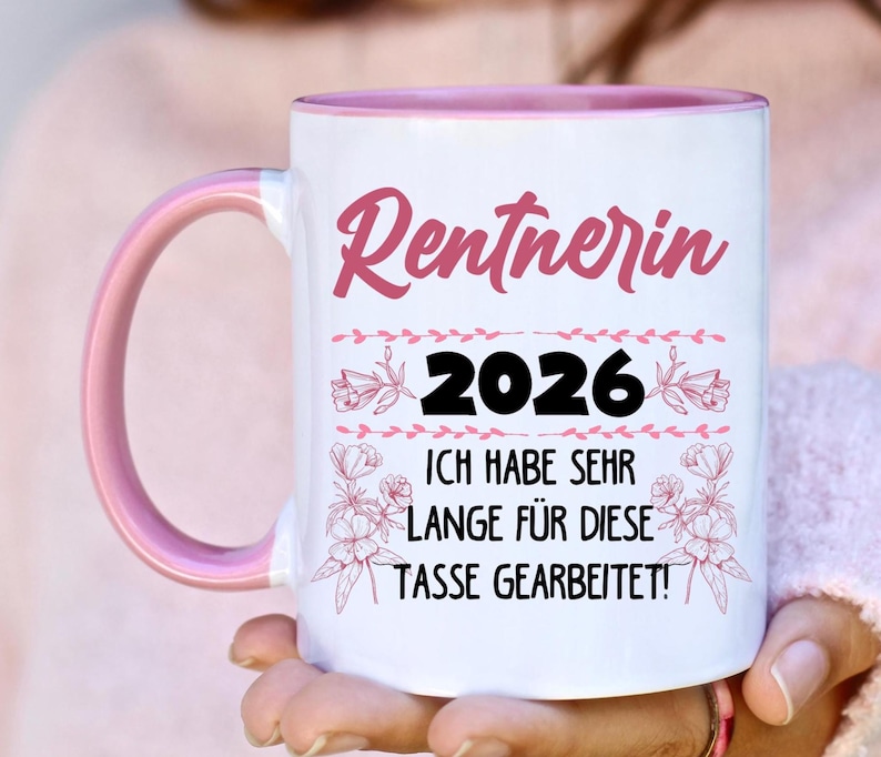 May include: White ceramic mug with a pink interior and handle. The mug features the text "Rentnerin 2026" in pink script, with additional text below. Floral accents surround the text.