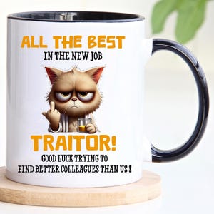 Op de afbeelding: Een witte keramische mok met een zwarte handgreep en rand. De mok heeft een cartoonkat met een chagrijnige uitdrukking die een glas sinaasappelsap vasthoudt. De tekst op de mok luidt "All the best in the new job Traitor! Good luck trying to find better colleagues than us!"