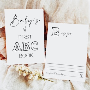 May include: Two white cards with black text. One reads "Baby's First ABC Book" and the other says "B is for". A small bouquet of white flowers rests near the cards. The background is a soft, neutral fabric.