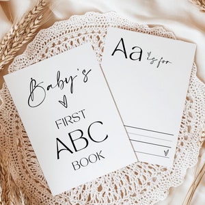 May include: A white card with the text "Baby's First ABC Book" and a heart symbol. A second white card with the text "Aa is for" and a heart symbol. Both cards are on a white crocheted doily with wheat stalks on a beige fabric background.