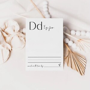 May include: A white card with black text that says "Dd is for" with a blank space to write in. The card also says "made with love by: " with a blank space to write in. The card is on a white surface with white pom poms and a brown fan.