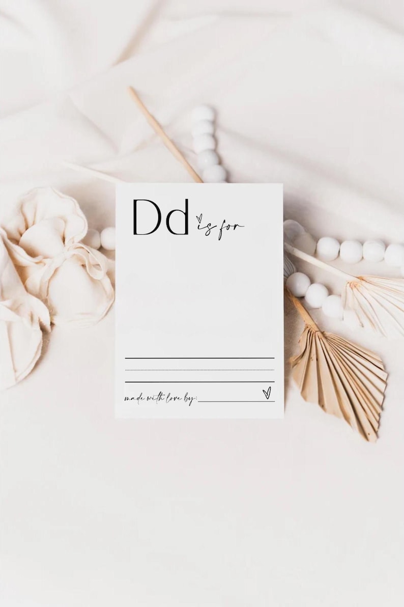 May include: A white card with black text that says "Dd is for" with a blank space to write in. The card also says "made with love by: " with a blank space to write in. The card is on a white surface with white pom poms and a brown fan.