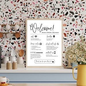 K&ouml;nnte beinhalten: Ein schwarz-wei&szlig;es Willkommensschild mit Informationen &uuml;ber WLAN, Standort, Hausordnung, Check-out-Informationen und Kontaktinformationen. Das Schild ist gerahmt und h&auml;ngt an einer Wand mit einem Terrazzo-Muster.