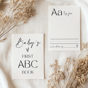 May include: A white card with the text "Baby's First ABC Book" in black. A second white card with the text "Aa is for" in black. Both cards are on a white fabric background with dried flowers.
