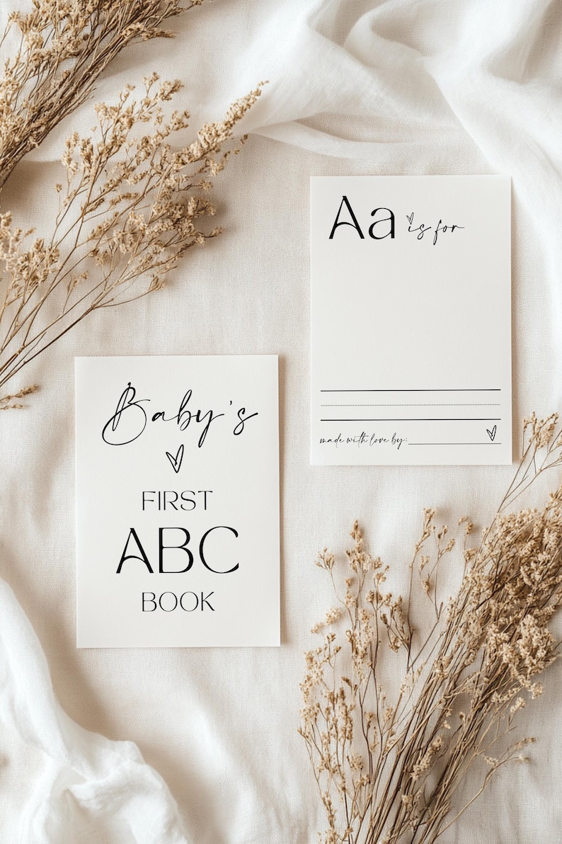 May include: A white card with the text "Baby's First ABC Book" in black. A second white card with the text "Aa is for" in black. Both cards are on a white fabric background with dried flowers.