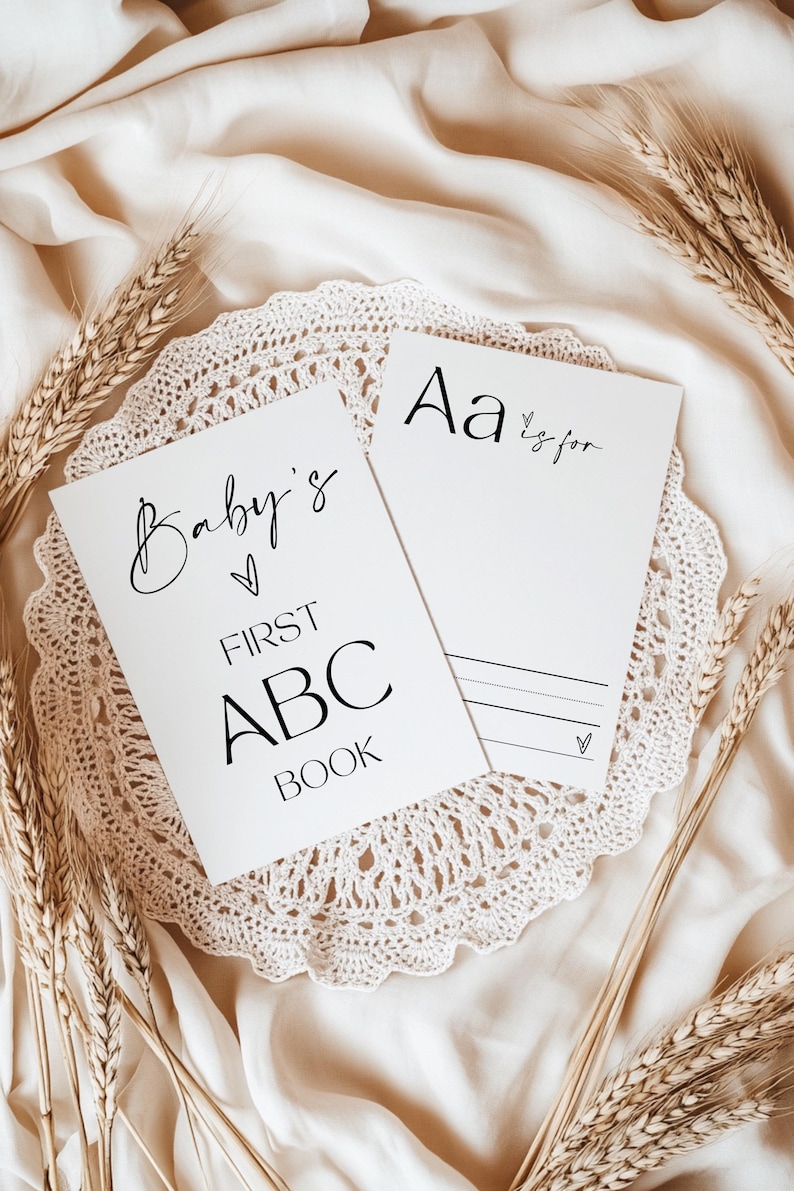 May include: A white card with the text "Baby's First ABC Book" and a heart symbol. A second white card with the text "Aa is for" and a heart symbol. Both cards are on a white crocheted doily with wheat stalks on a beige fabric background.