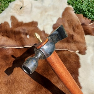 Op de afbeelding: Een hamer met een houten steel, een messing kop en een zwarte metalen kop. De hamer ligt op een bruin en wit koeienhuid.
