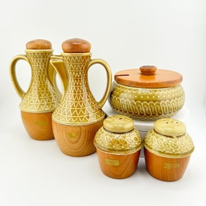 May include: A set of six vintage ceramic kitchenware pieces in a brown and green colour scheme. The set includes two oil and vinegar bottles, a salt and pepper shaker, and a covered dish with a wooden lid.