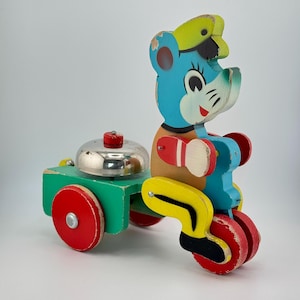 May include: Vintage wooden toy featuring a blue bear character riding a tricycle pulling a green cart with a silver bell. The toy has red, yellow, and black accents, showcasing a classic, nostalgic design. The toy is likely from the mid-20th century.