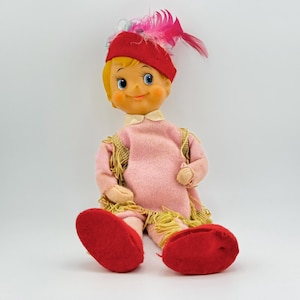 Puede incluir: Una muñeca de elfo vintage con cabello rubio, vestida con un vestido rosa con flecos dorados, un sombrero rojo con una pluma y zapatos rojos.
