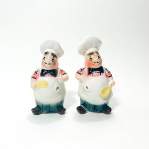 Pode incluir: Um par de saleiros e pimenteiros de cerâmica em forma de chefs. Os saleiros usam chapéus de chef brancos, camisas cor-de-rosa, gravatas borboleta pretas e aventais brancos. O saleiro segura uma espátula amarela e o pimenteiro segura uma colher amarela. Os saleiros estão em pé sobre uma superfície branca.